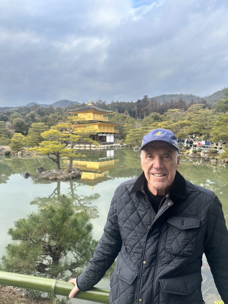 A man smiles in front of the Golden Pavilion in Kyoto, reflecting in the tranquil pond surrounded by lush greenery.