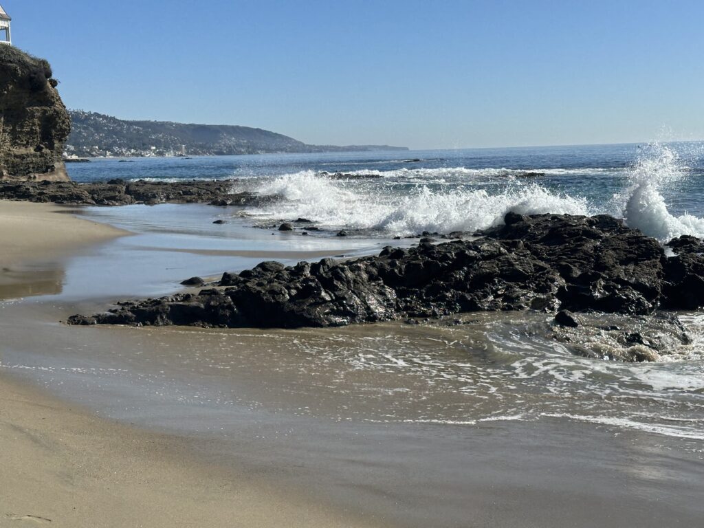 Calm beach scene with gentle waves lapping against rocky shore, framed by distant hills and clear blue sky, capturing a peaceful coastal vibe.