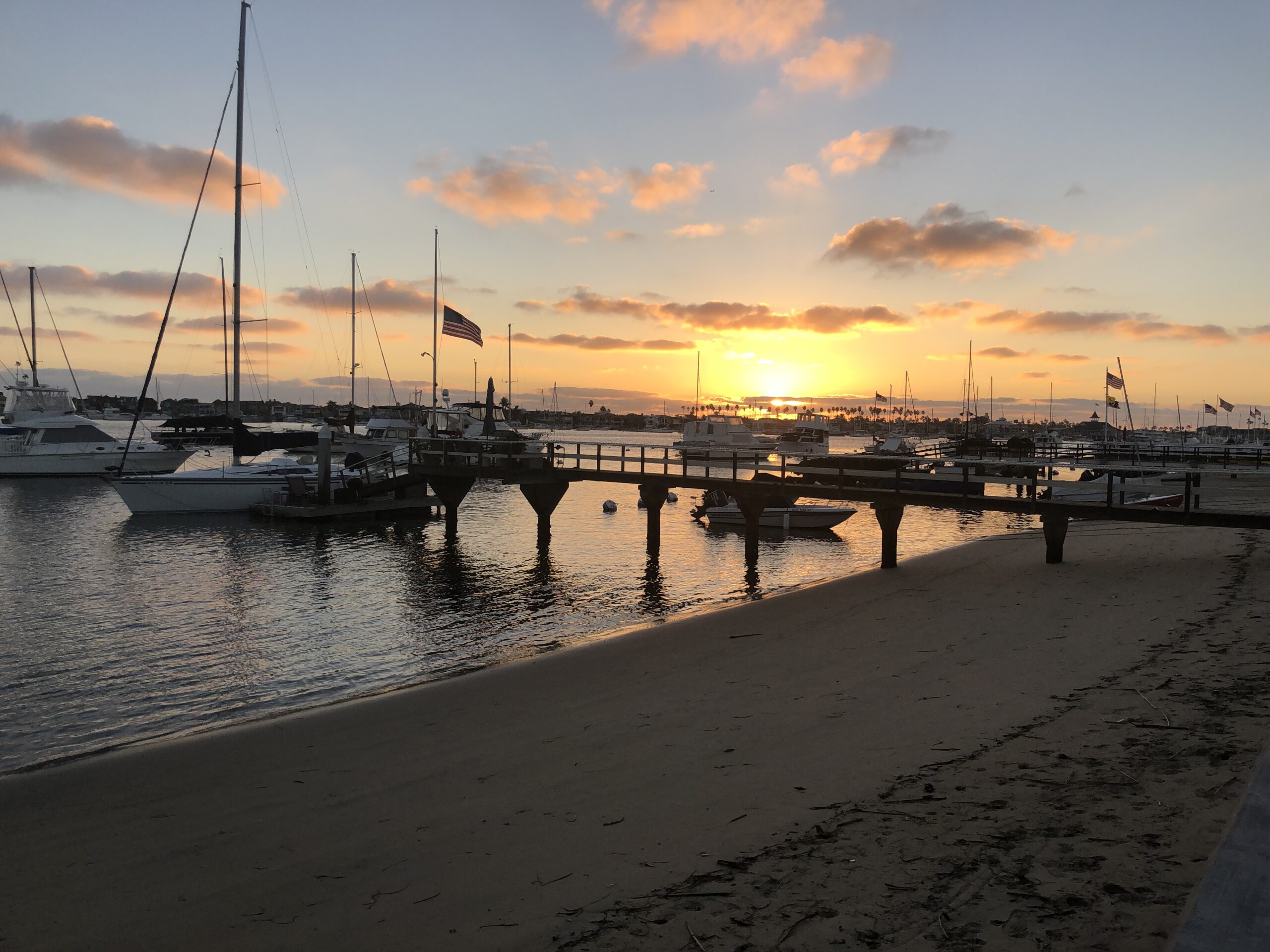 Sunset over a marina with sailboats and motorboats, reflecting on calm waters. An American flag flutters on a nearby dock, enhancing the serene atmosphere.