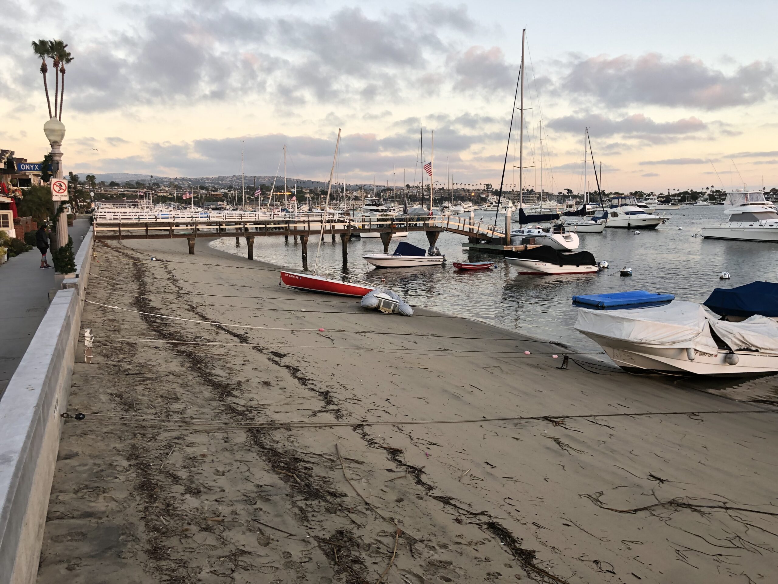 Sunset view of a marina with moored boats and a sandy shore, featuring a boardwalk lined with palm trees and distant hills.